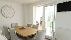 Une salle à manger avec une table en bois, six chaises rembourrées, une horloge murale et une porte-fenêtre coulissante donnant sur l’extérieur.