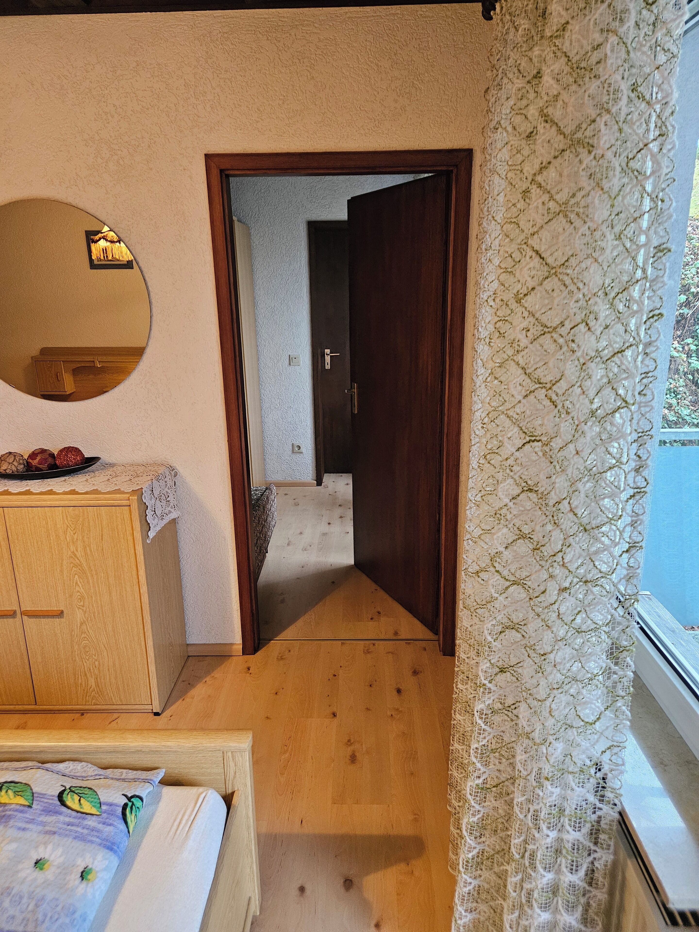 Ein Schlafzimmer mit einem Holzkoffer, einem Bett mit weißer Bettdecke und einem Fenster mit Spitzenvorhängen.