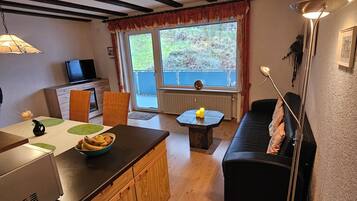 Un espace de vie chaleureux avec un plancher en bois, un canapé noir, une table à manger et un coin cuisine équipé d’une télévision et d’une lampe.