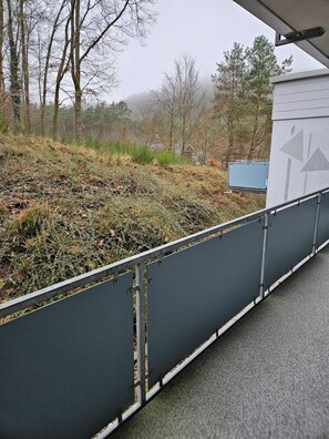 Et hegn i metal med en betonfundament, en græsklædt skråning og en bygning med en grå facade i baggrunden.