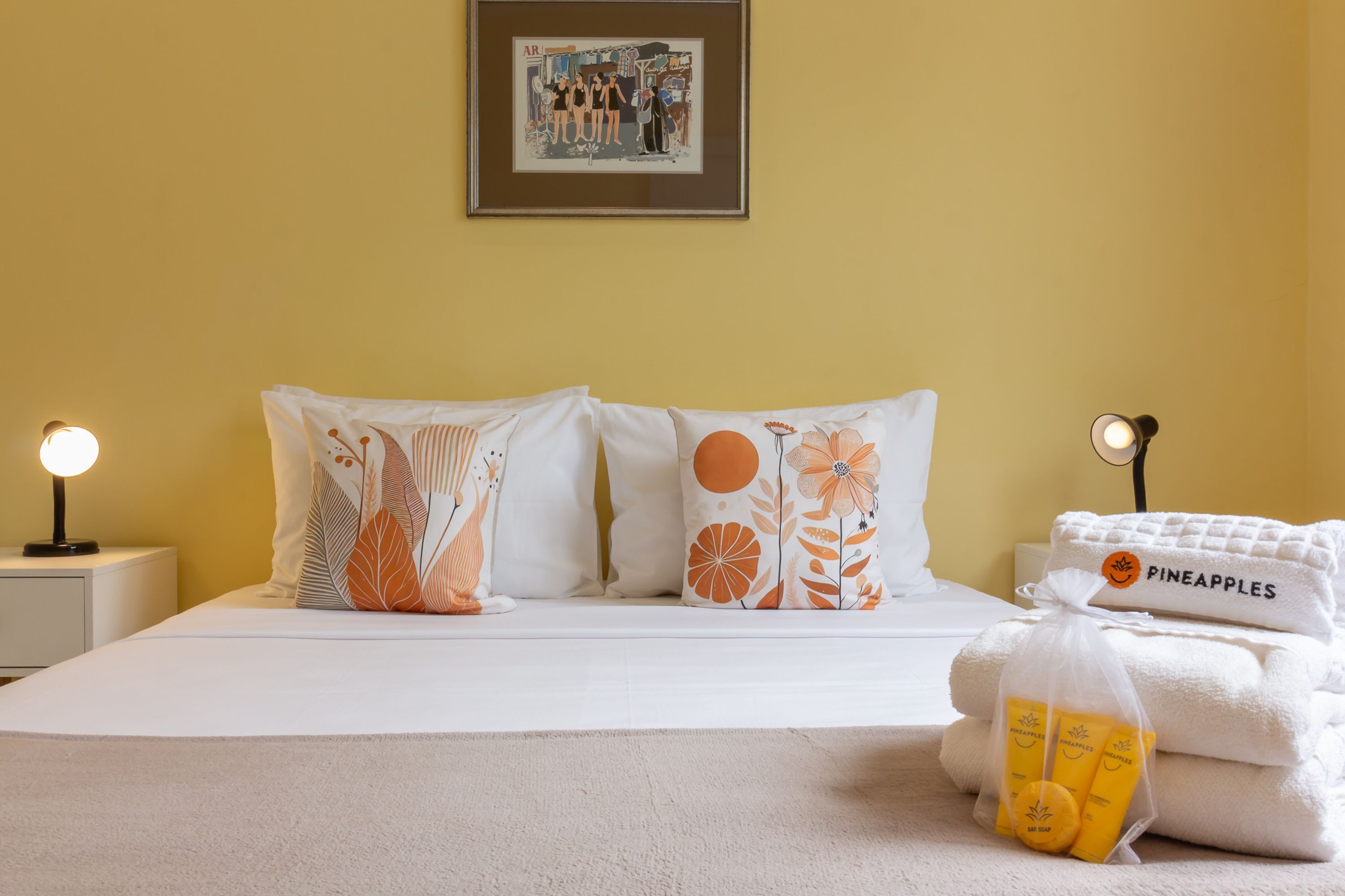 A neatly made bed with floral-patterned pillows, a framed picture on the wall, and a small bedside table with a lamp.