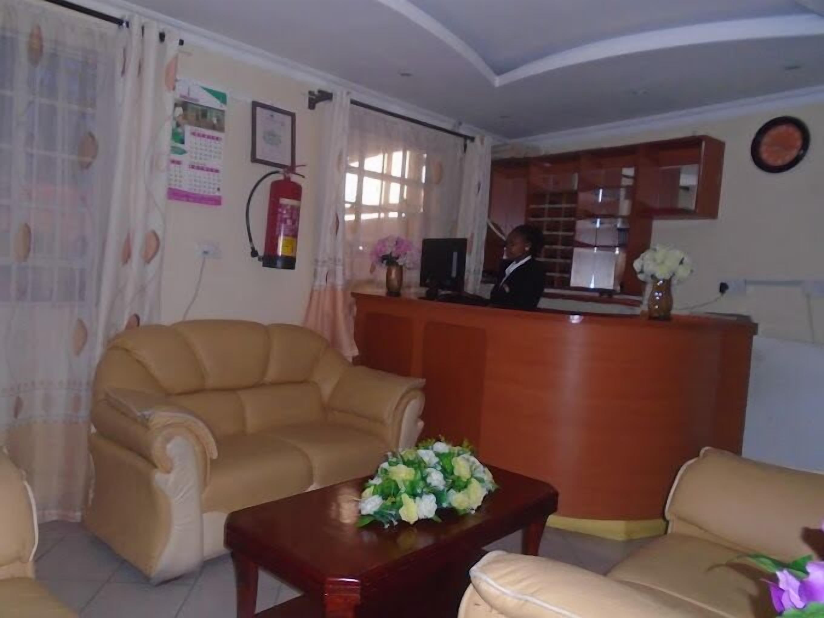 A hotel lobby with a reception desk, two sofas, a coffee table with flowers, a fire extinguisher, and a calendar on the wall.