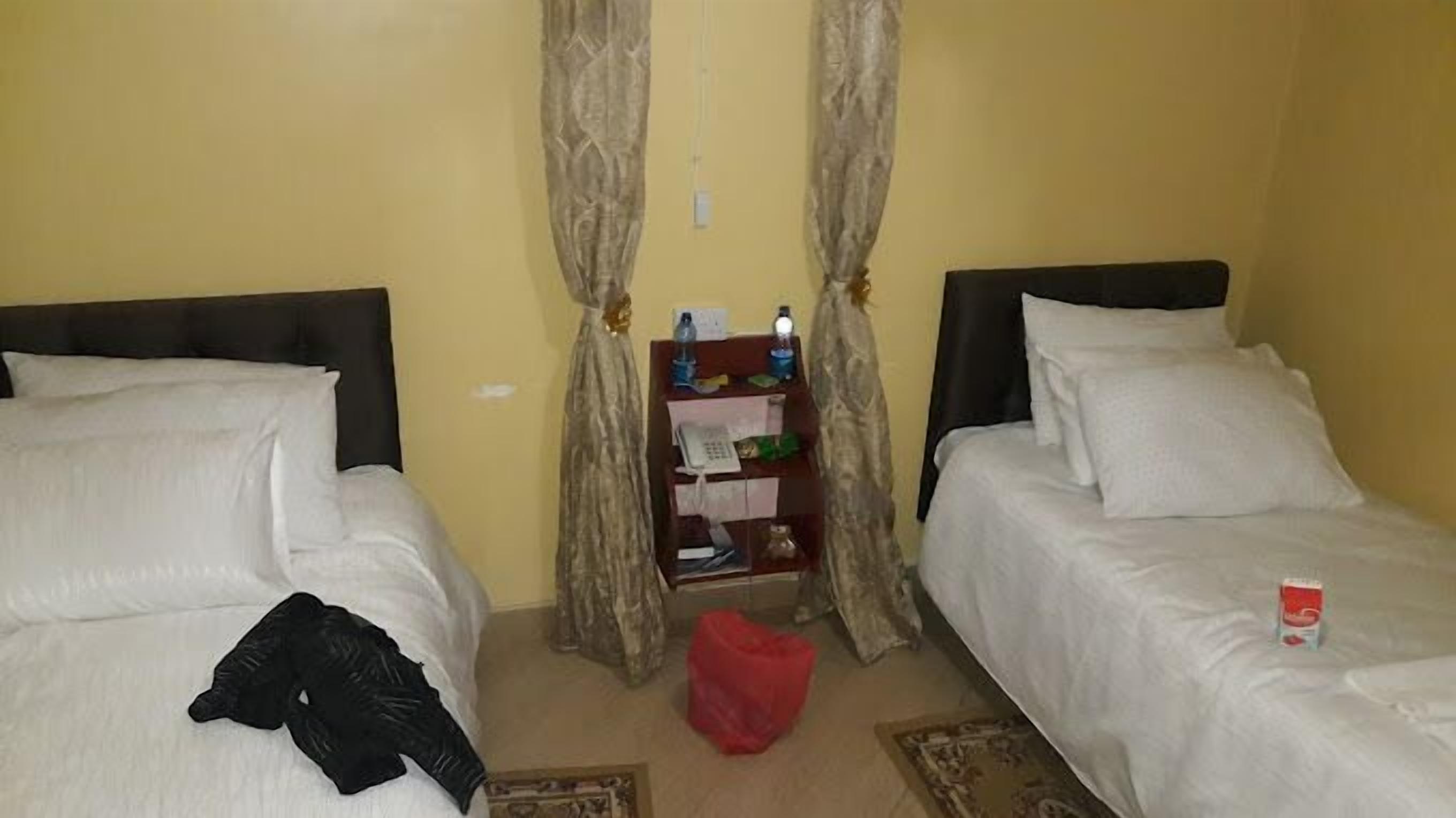 A room with two beds, a small shelf with personal items, and a red bag on the floor.