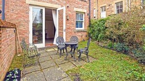 A brick house with a patio area featuring a table and chairs, a sliding glass door, and a small garden. - Churchside (Alnwick)