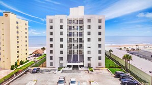 A multi-story hotel with a central entrance, parking lot, and a view of the beach.