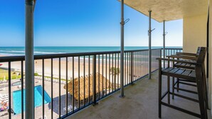 A balcony with a view of the beach and ocean, metal railings, and a pool area with lounge chairs.