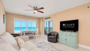 A living room with a sofa, a television, a ceiling fan, a dining area with a table and chairs, and a view of the ocean.
