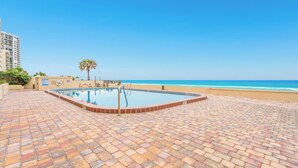 A swimming pool with a tiled deck, a palm tree, and a beach in the background. - Oceanfront | No-drive Beach | Sea Fern #303 (Daytona Beach Shores)