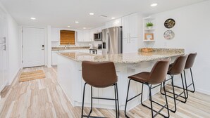 A modern kitchen with white cabinets, a granite countertop, stainless steel appliances, and bar stools. - Oceanfront | No-drive Beach | Sea Fern #303 (Daytona Beach Shores)