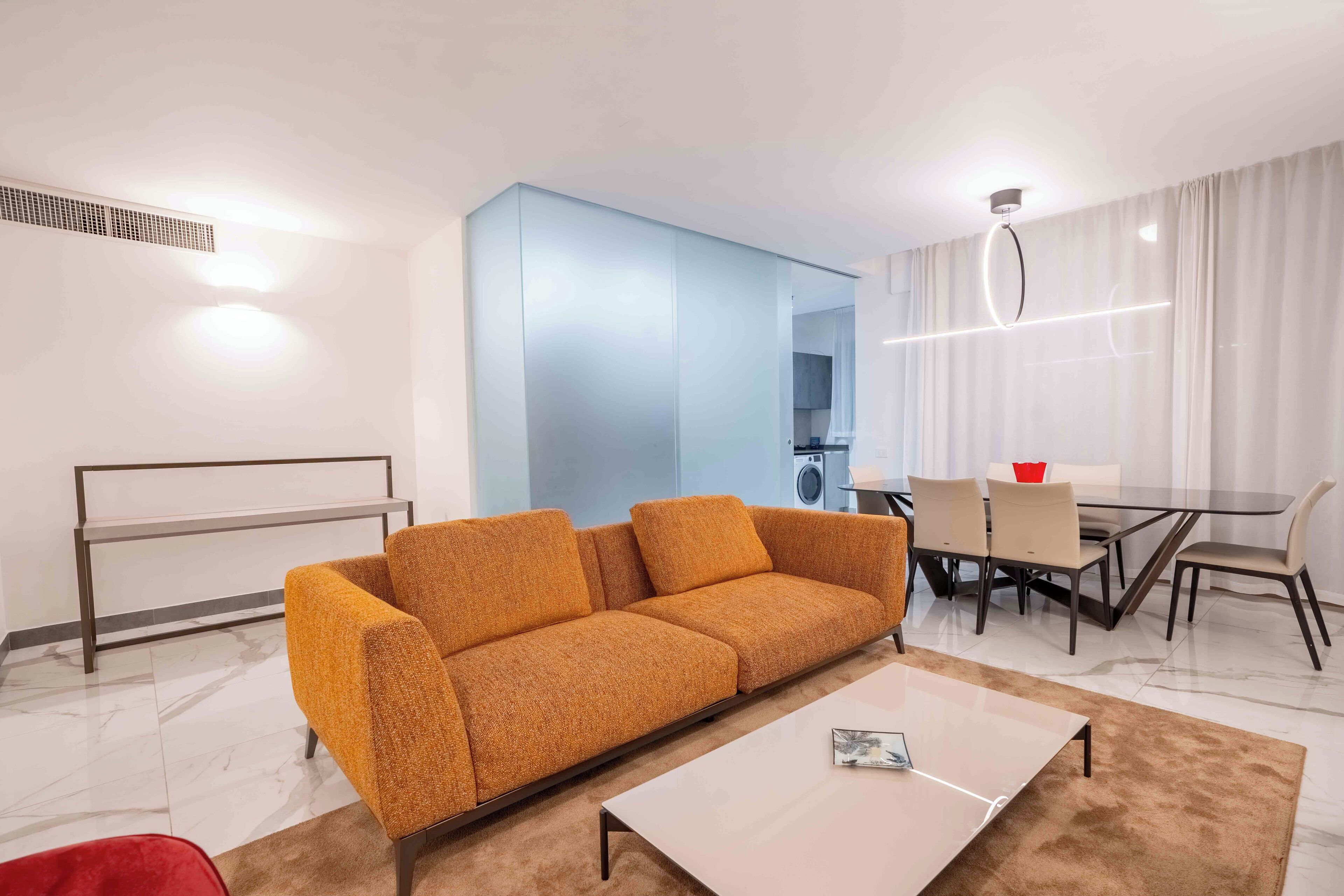 A modern living room with a sofa, a coffee table, a dining table with chairs, and a kitchen area in the background.