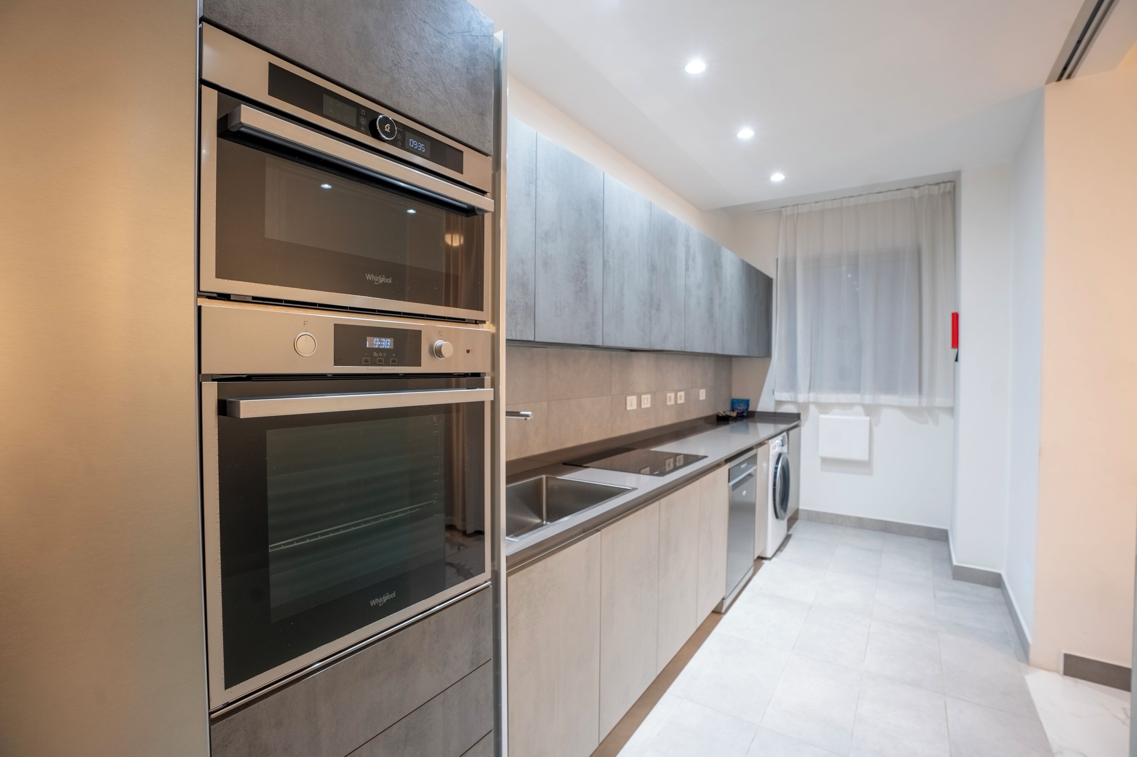 A modern kitchen with stainless steel appliances, a built-in oven, a microwave, a dishwasher, and a large window with curtains.