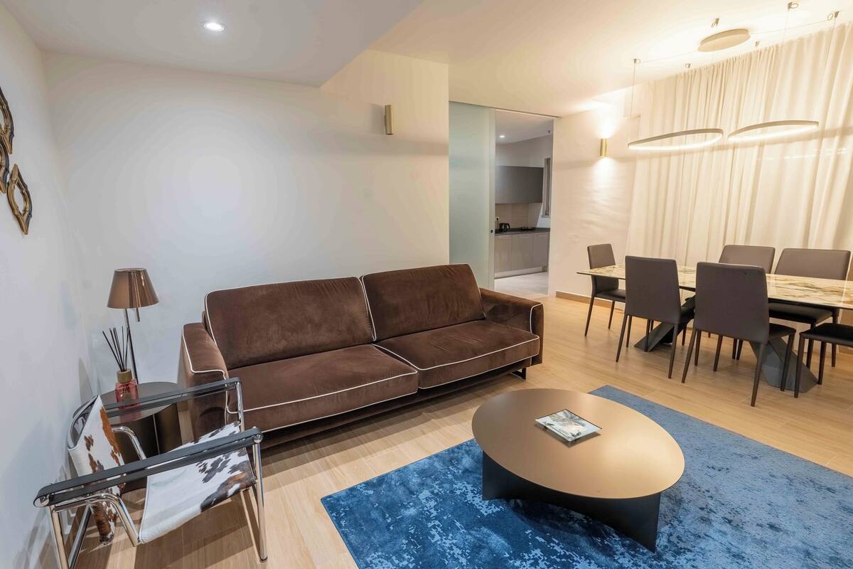 Een moderne woonkamer met een bruine bank, een glazen salontafel, en een eethoek met een tafel en stoelen.