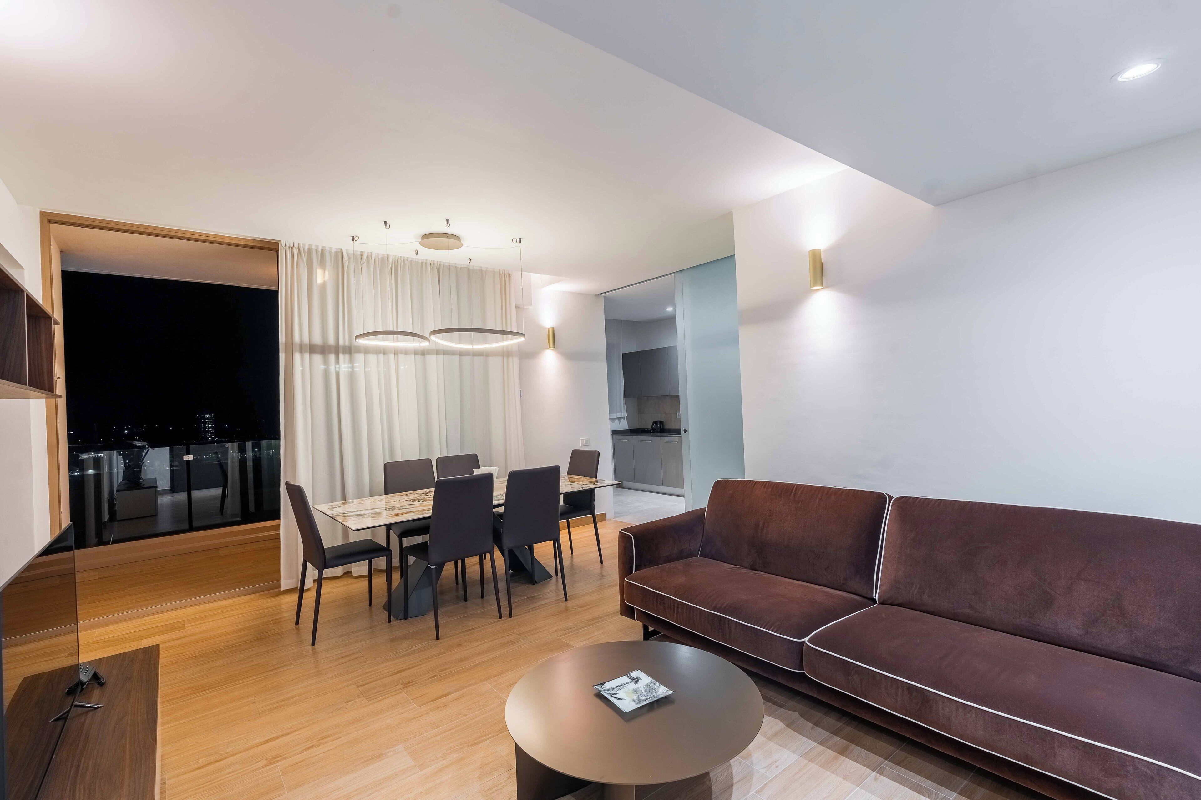 A modern living room with a brown sofa, a dining table with chairs, a coffee table, and a view of the city at night through a glass door.