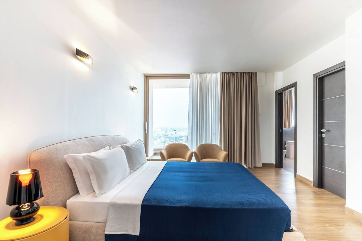 Een hotelkamer met een bed, een nachtkastje met lamp, twee stoelen, een groot raam met gordijnen en uitzicht op de stad.
