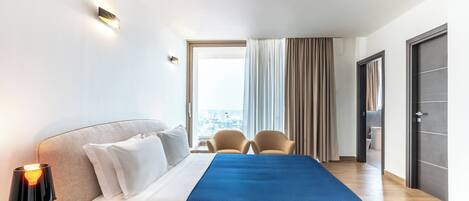 A hotel room with a bed, a nightstand with a lamp, two chairs, a large window with curtains, and a view of the city.