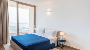 Habitación de hotel con una cama grande, una mesita de noche, una lámpara y vista a un cuerpo de agua a través de una puerta corrediza de vidrio.