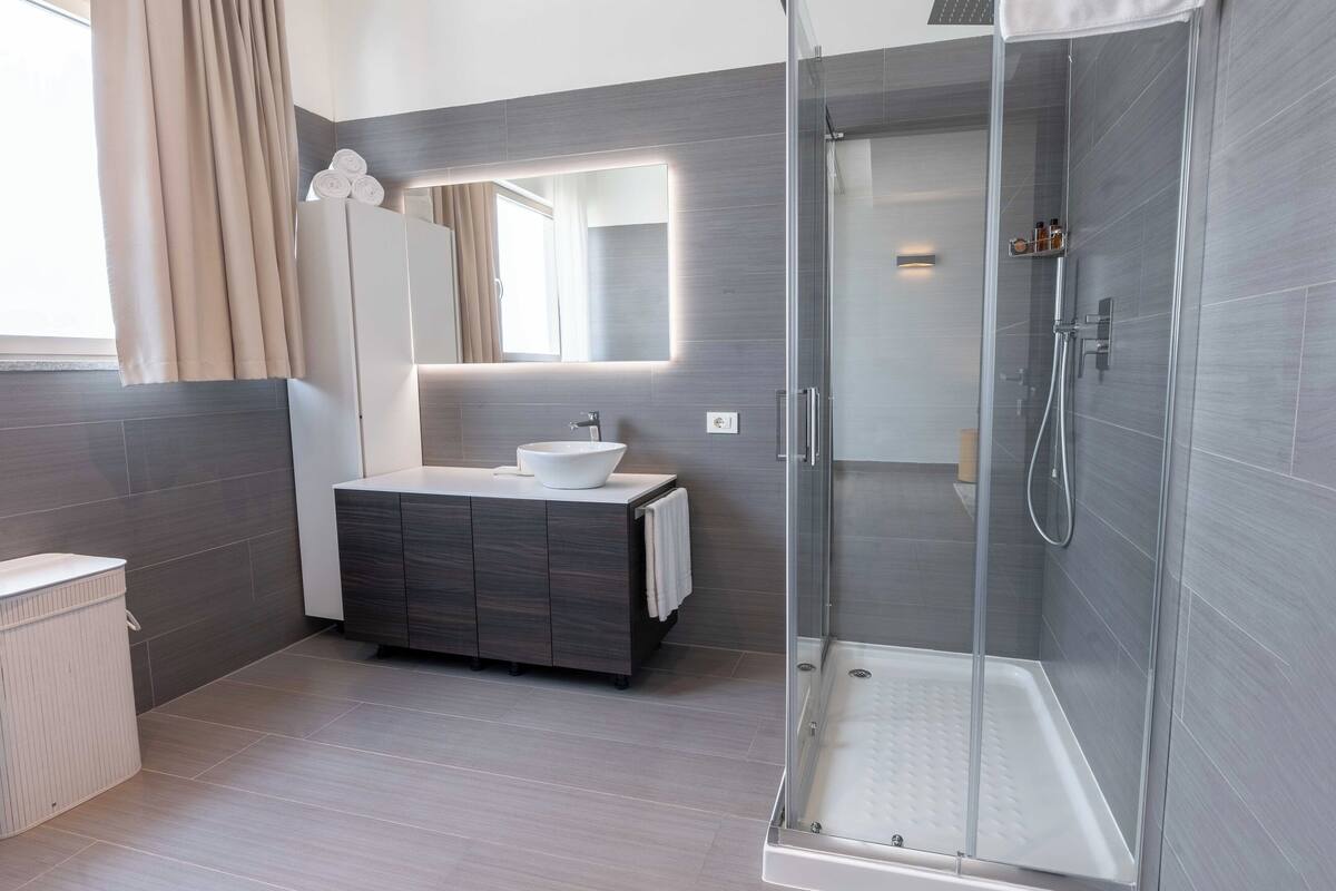 Een moderne badkamer met een douchecabine van glas, een wit wastafeltje en een donkere badkamerkast.