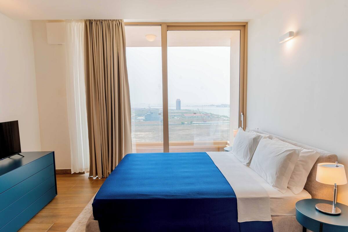 Een hotelkamer met een bed, een televisie, een bureau, een lamp en uitzicht op een stadsgezicht door grote ramen.
