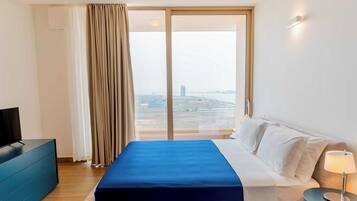 Habitación de hotel con cama, televisión, escritorio, lámpara y vistas de la ciudad a través de amplios ventanales.