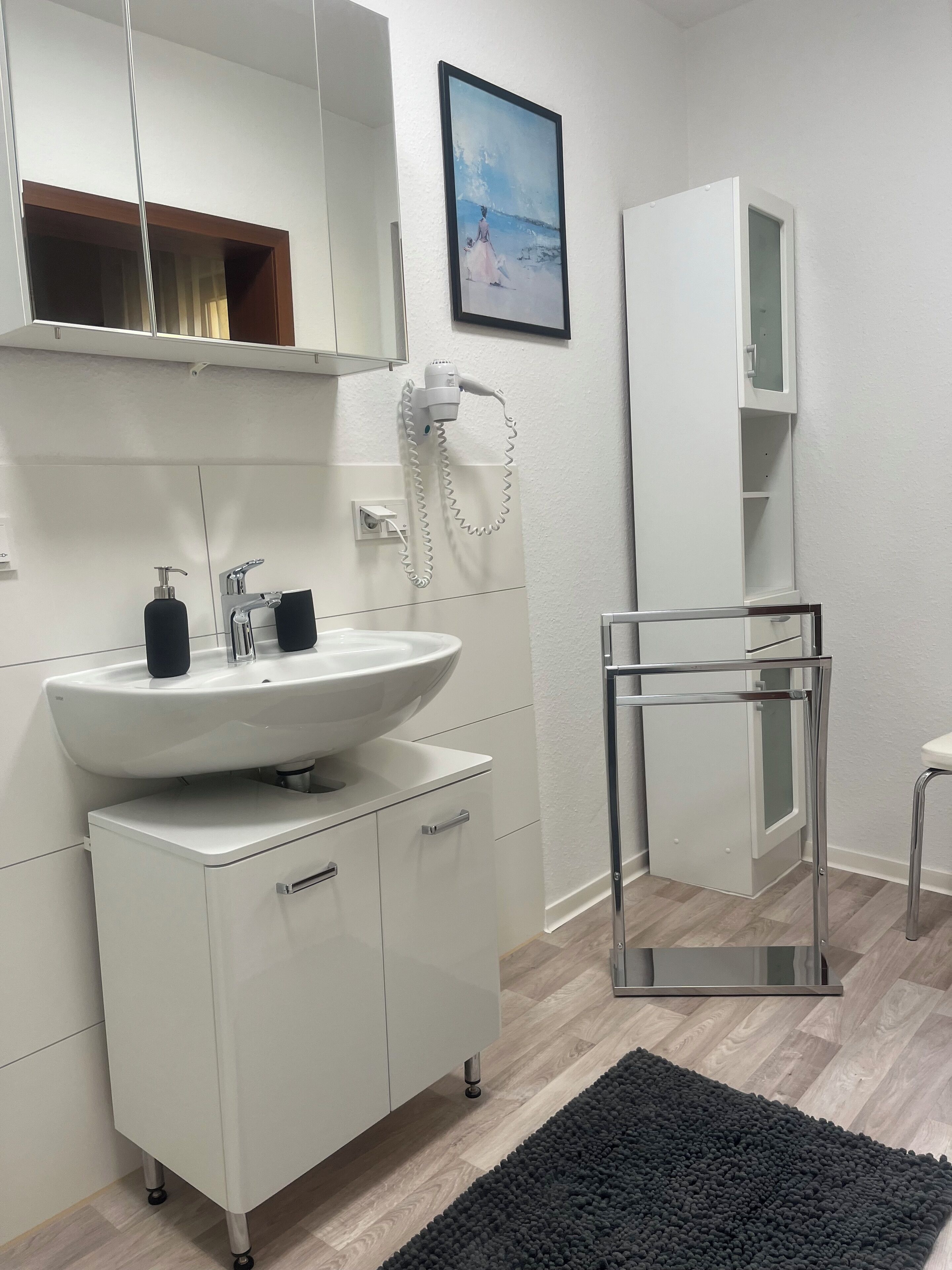 Une salle de bain moderne avec un lavabo blanc, un miroir et un meuble, un sèche-cheveux et un tapis noir sur le sol.