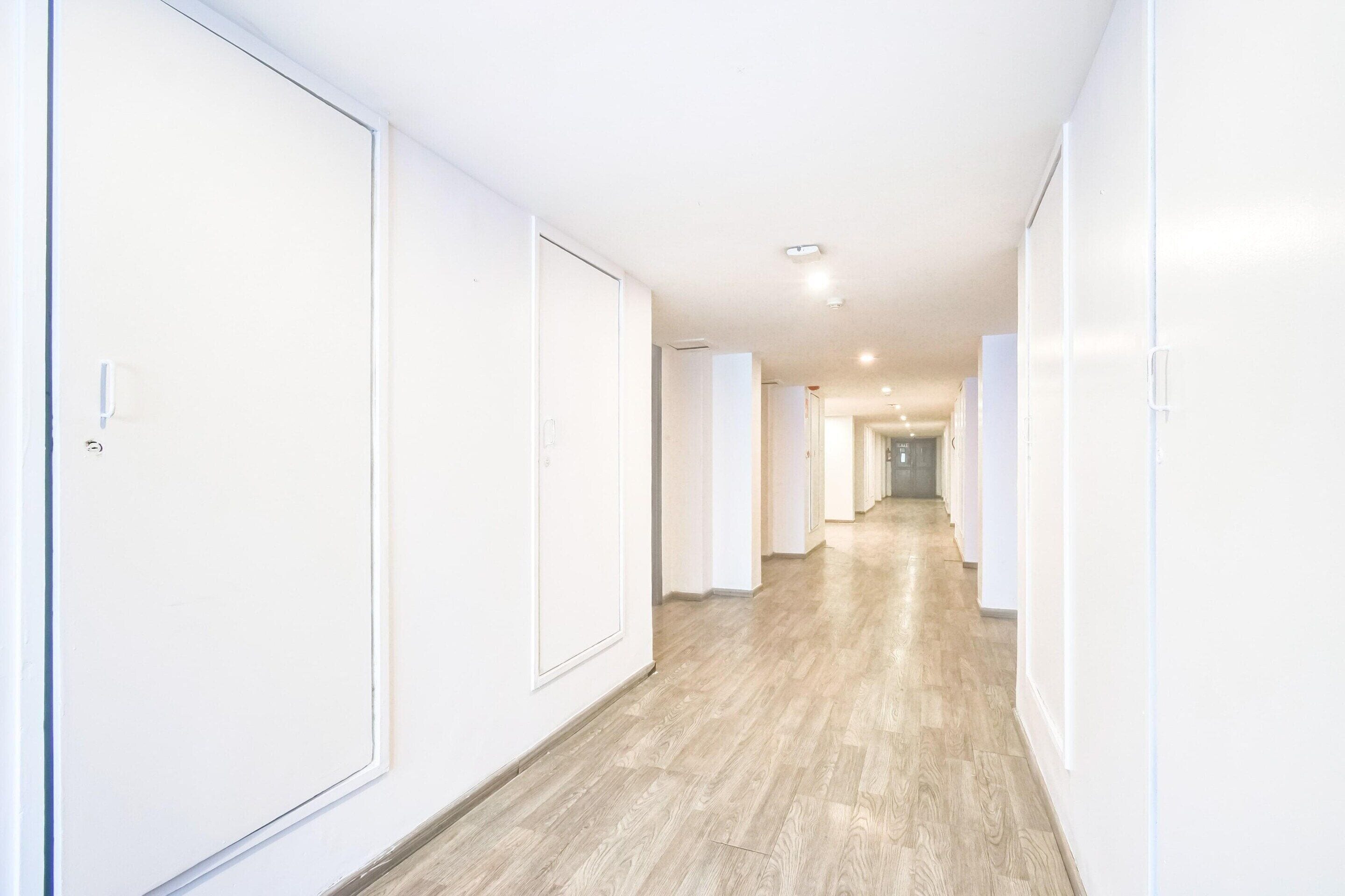 A hallway with white walls and wooden flooring, leading to a door at the end.
