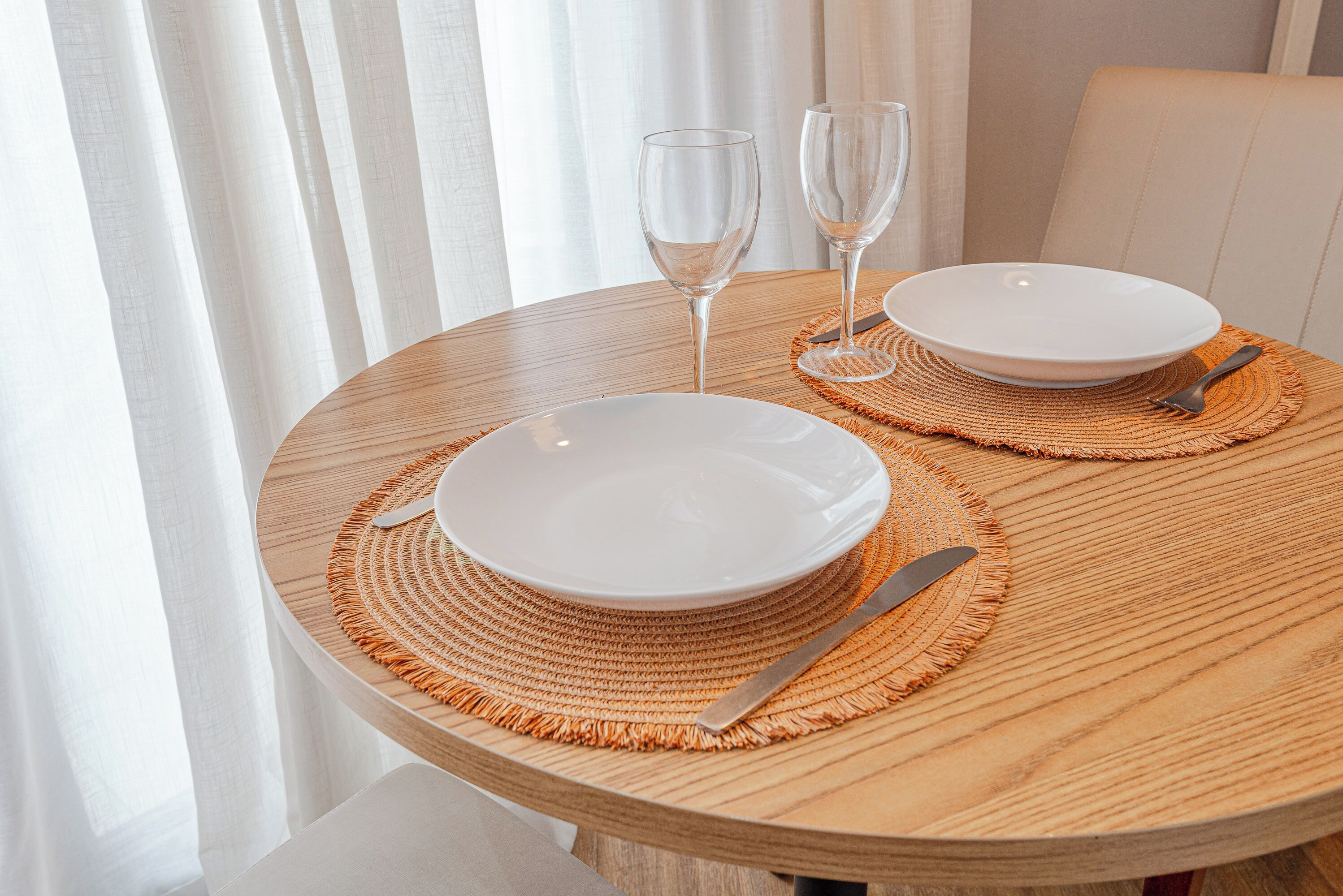A round wooden table with two white plates, a knife, and two wine glasses, set on a woven placemat.
