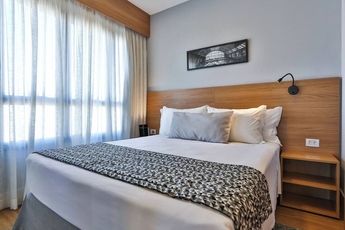Hotellihuone, jossa on suuri sänky, sängynpääty, jossa on valaisin, yöpöytä ja seinälle ripustettu taulu.