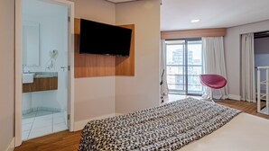 Une chambre d’hôtel moderne dotée d’un grand lit, d’un téléviseur fixé au mur, d’une salle de bain avec une porte en verre et offrant une vue sur la ville.