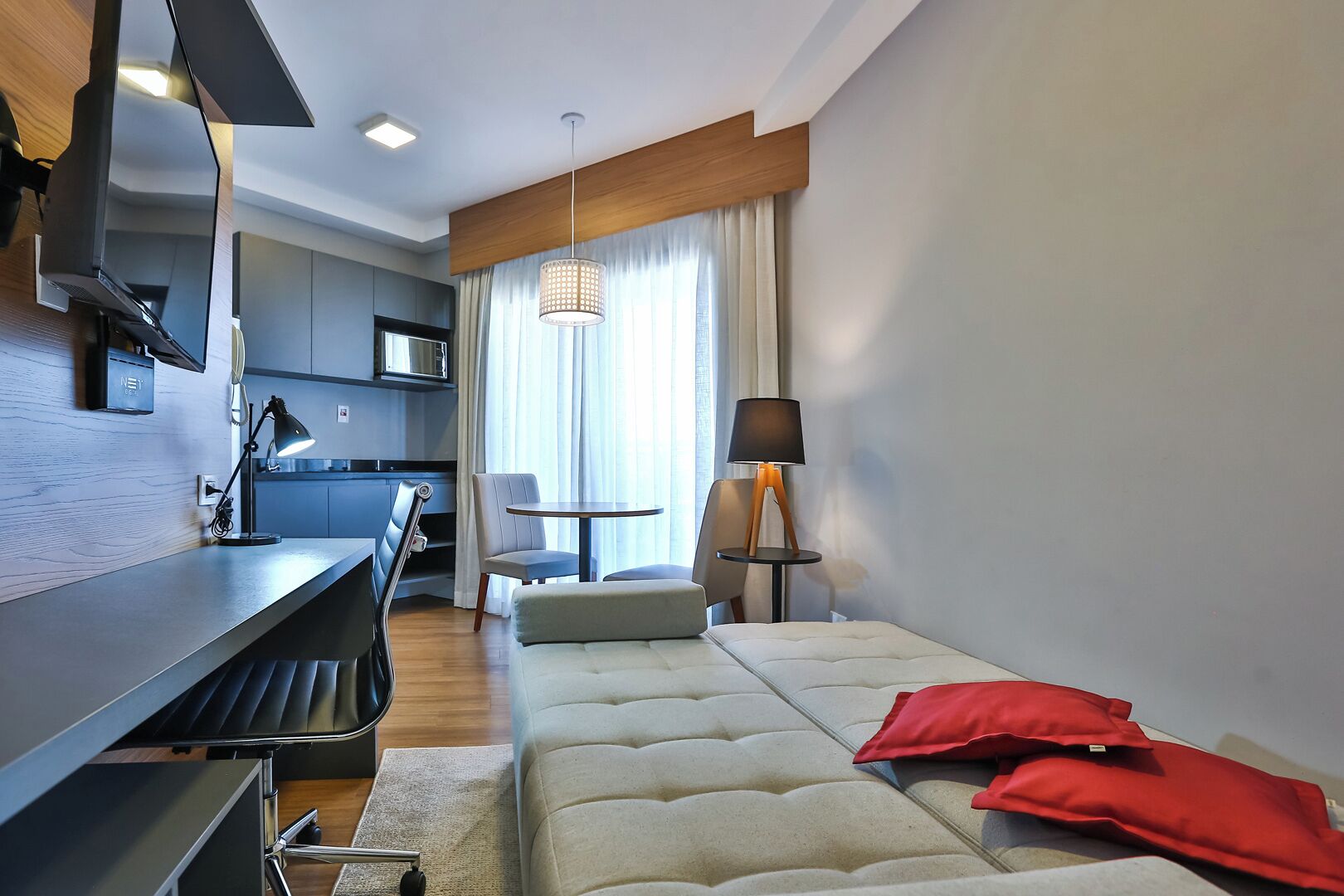 A modern hotel room with a kitchenette, a dining area, and a bed with red pillows.