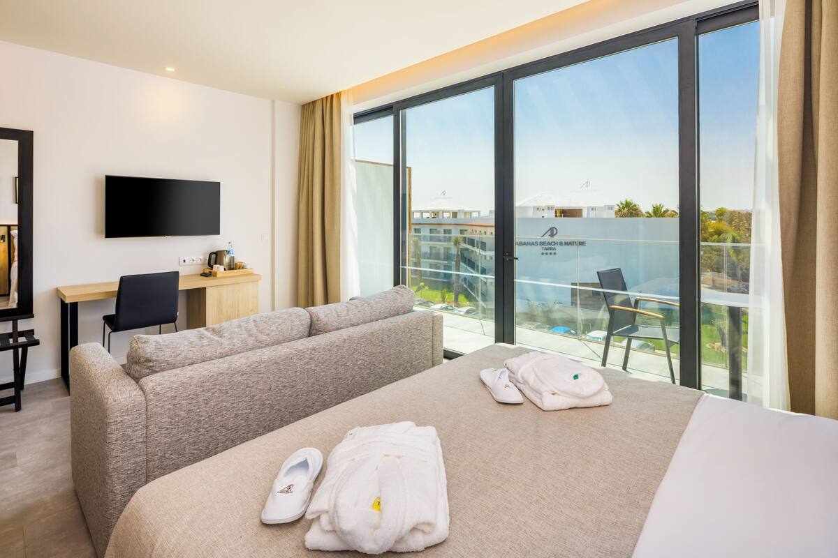 Habitación de hotel con cama, sofá, escritorio, televisor y balcón con vistas.