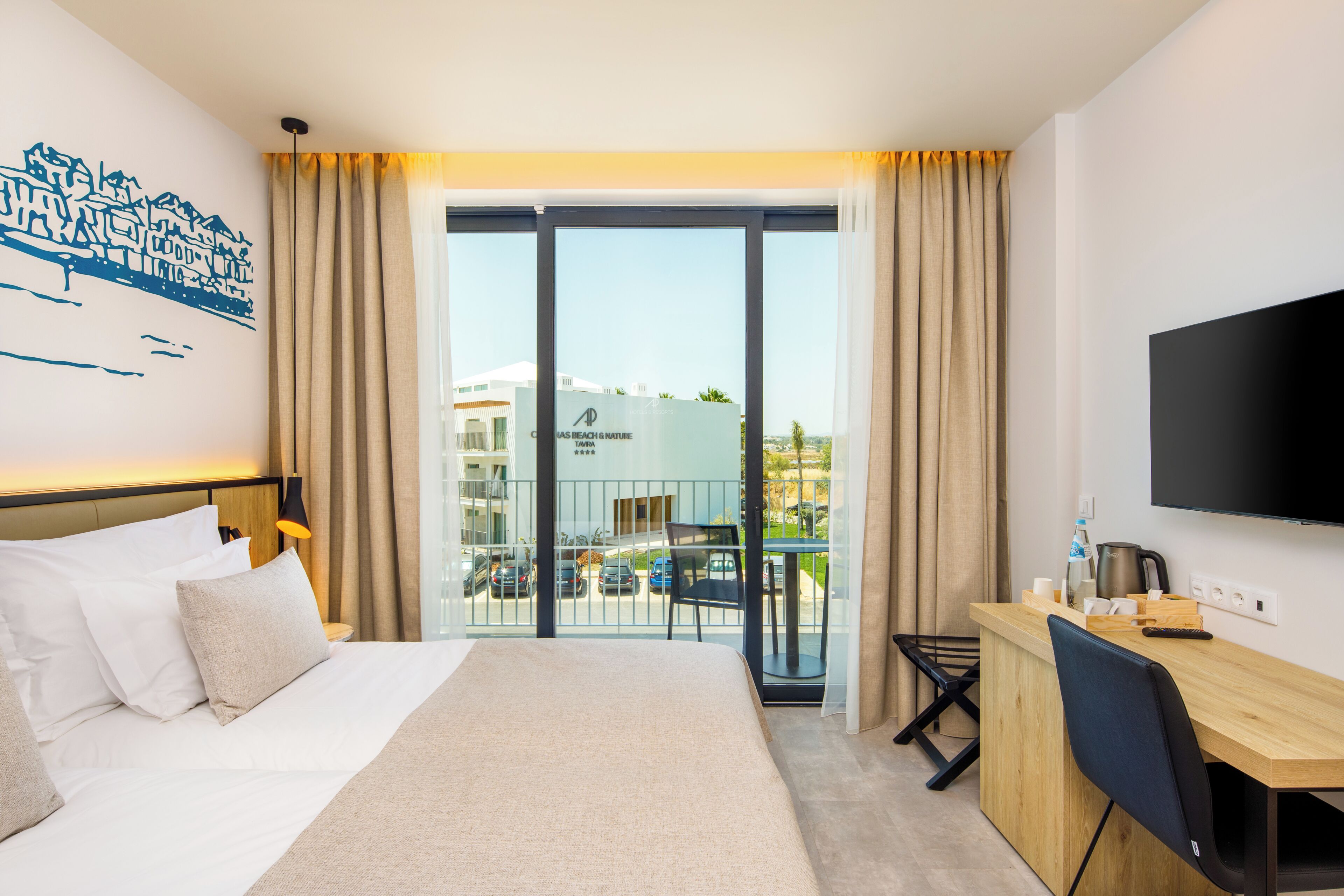 Ein Hotelzimmer mit Bett, Schreibtisch, Fernseher und einem Balkon mit Blick auf die Stadt.
