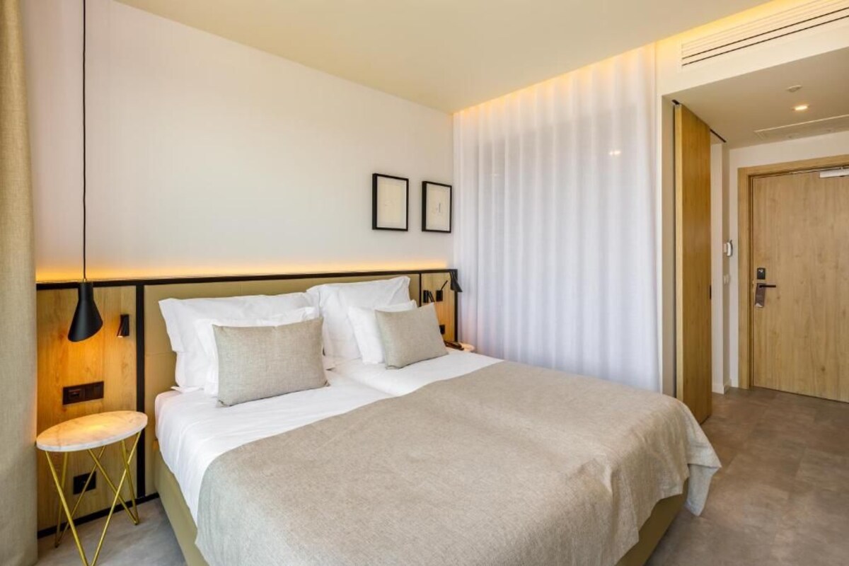 A modern hotel room with a large bed, bedside tables, a wall-mounted lamp, and a door.