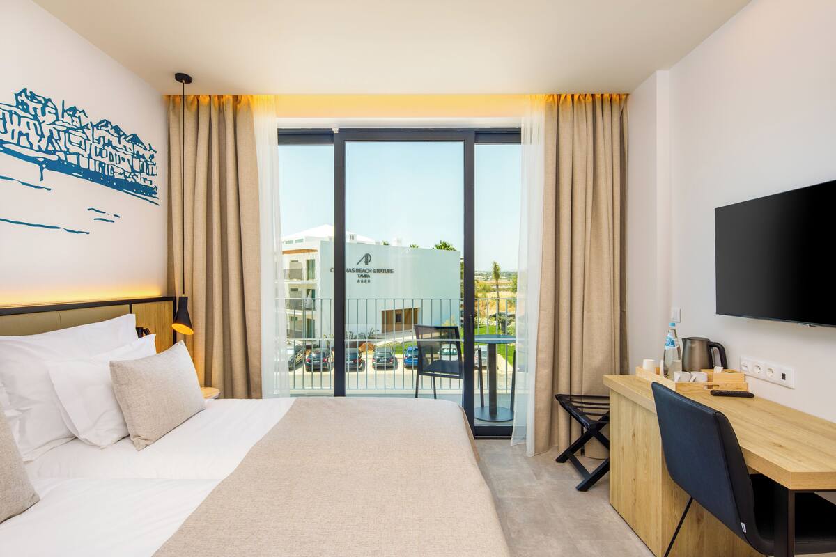 Habitación de hotel con cama, escritorio, televisor y balcón con vistas a la ciudad.