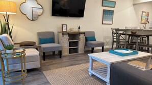 A modern living room with a flat-screen TV, two grey armchairs with teal cushions, a coffee table, and a dining area with a wooden table and chairs. - Charming 2 bedroom townhome in Seagrove steps to the beach! (Santa Rosa Beach)