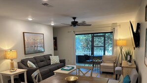 A modern living room with a dark brown sofa, a wooden coffee table, and a dining table with chairs. There is a ceiling fan, a floor lamp, and a TV mounted on the wall. - Charming 2 bedroom townhome in Seagrove steps to the beach! (Santa Rosa Beach)