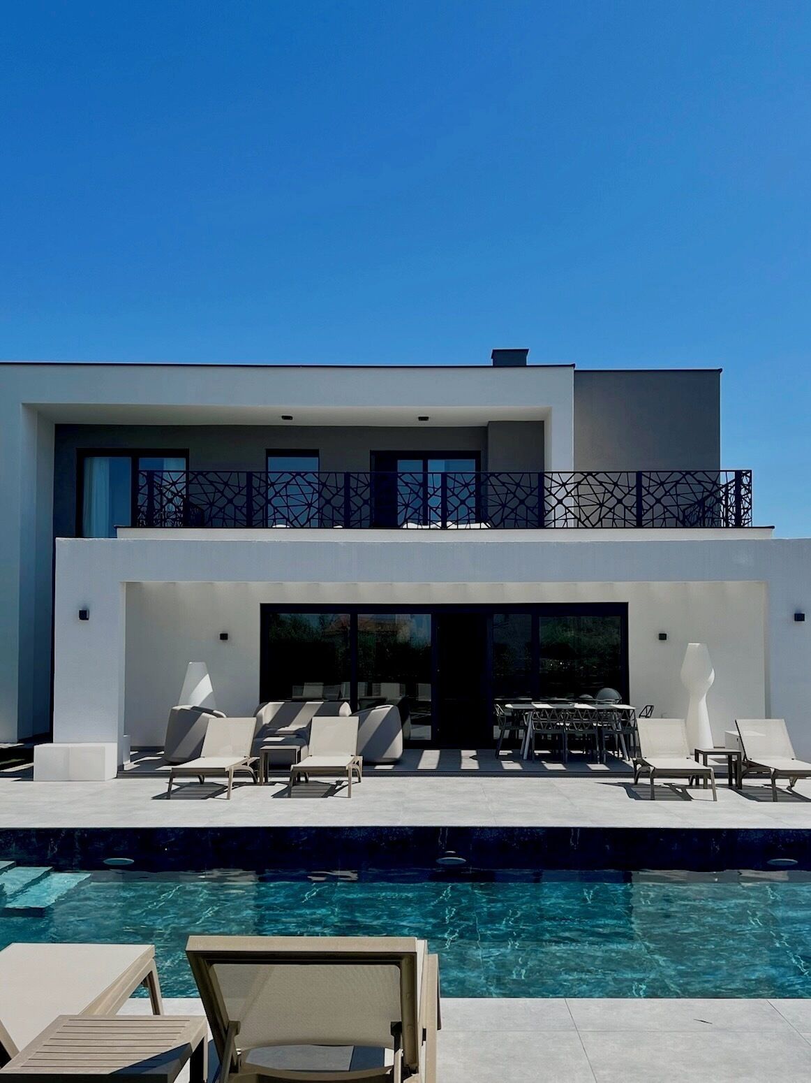 Ett modernt hus i två våningar med pool, utomhusmöbler och en balkong.