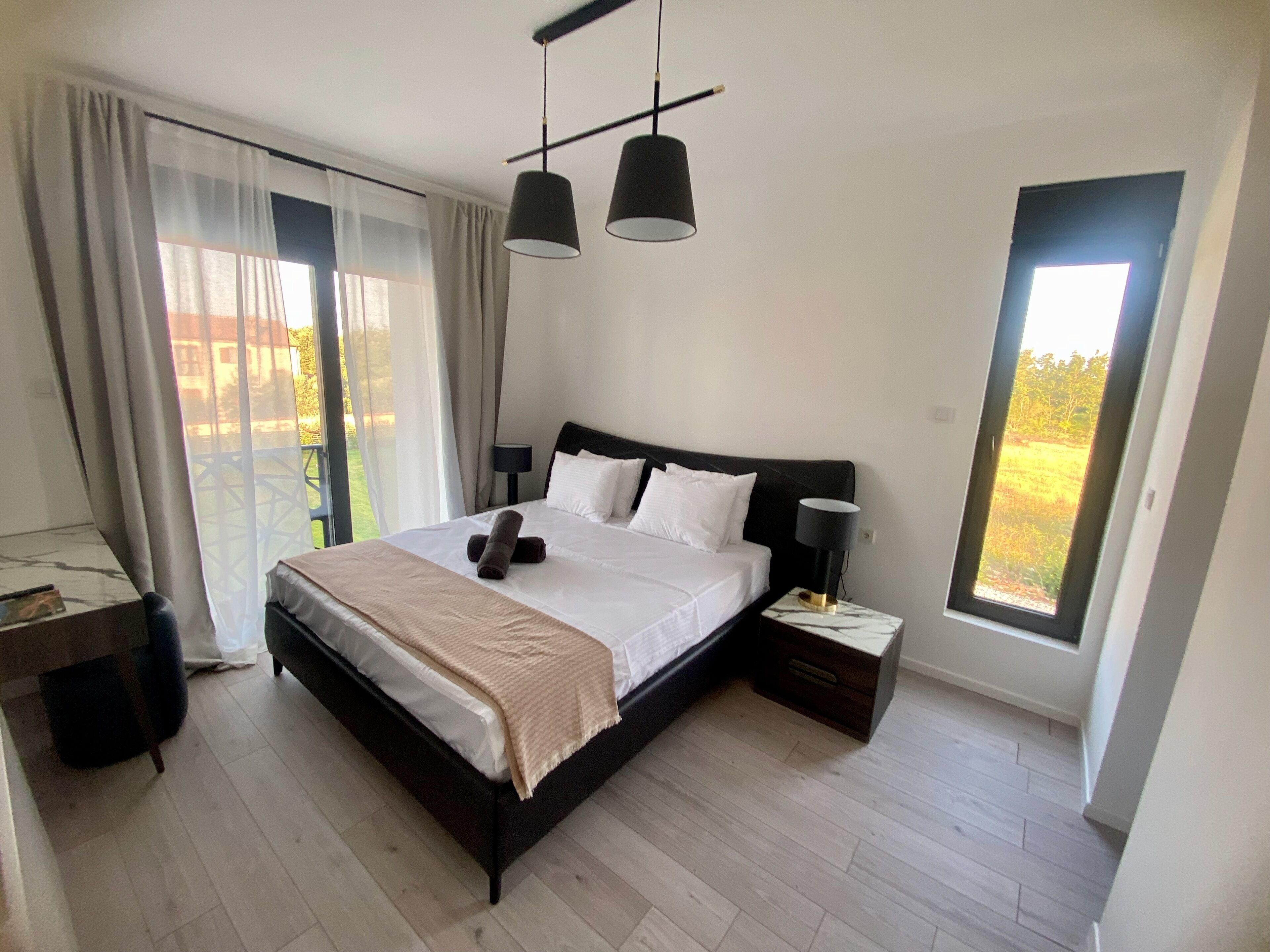 Ett modernt sovrum med en stor säng, två sänglampor, ett nattduksbord och utsikt över en trädgård genom ett fönster.
