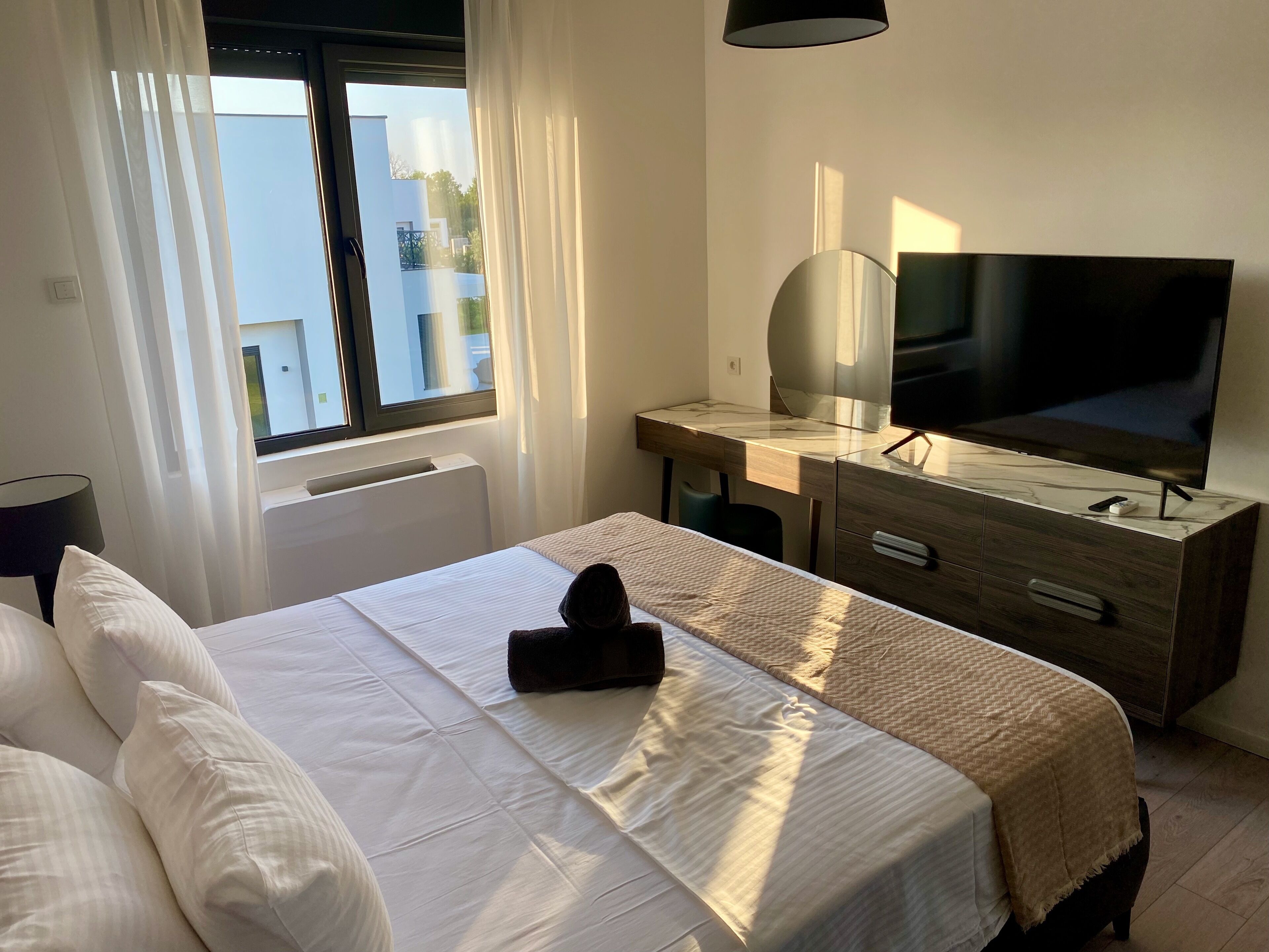 A hotel room with a bed, a desk with a TV, a window with curtains, and a radiator.