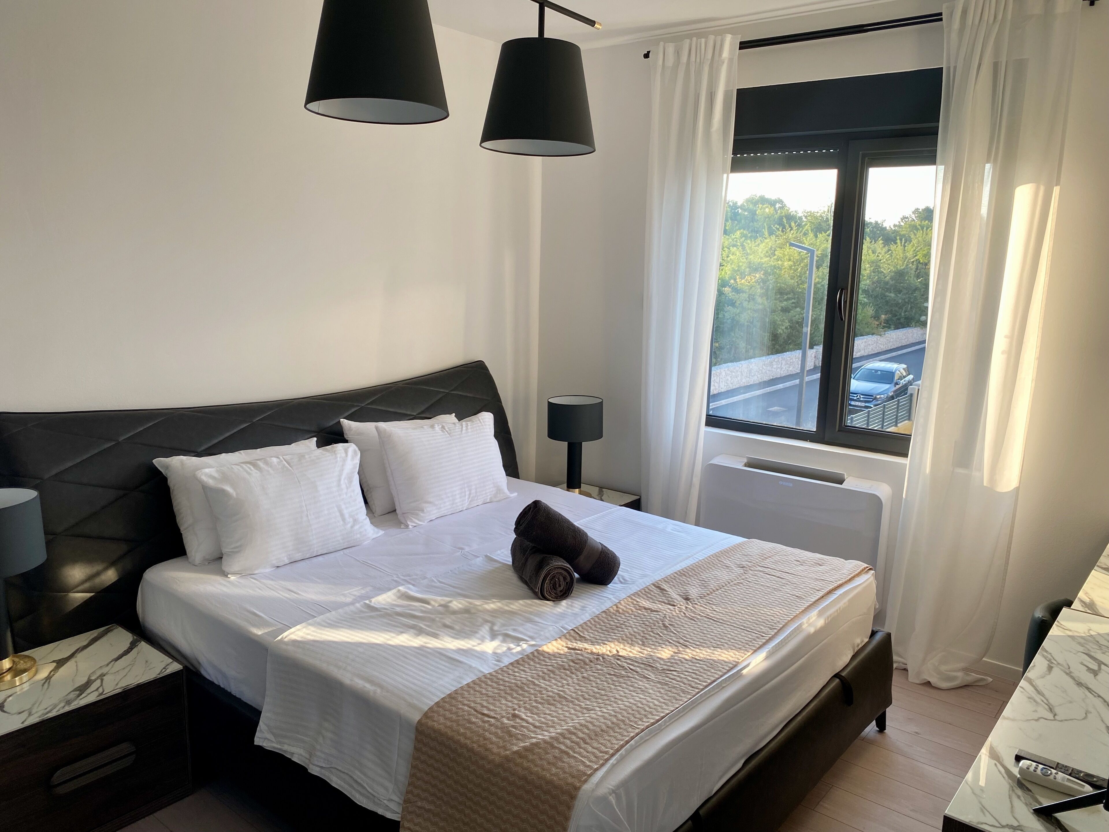 Una cama bien hecha con sábanas blancas y una toalla enrollada, un cabecero negro, una mesita de noche con una lámpara y una ventana con cortinas.