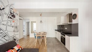 A modern kitchen with a black countertop and stainless steel appliances, a dining area with a wooden table and chairs, and a living area with a sofa and colorful cushions.