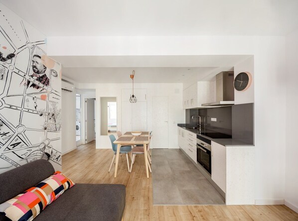 A modern kitchen with a black countertop and stainless steel appliances, a dining area with a wooden table and chairs, and a living area with a sofa and colorful cushions. - GuestReady – Your Cozy Escape in Valencia (Valencia)