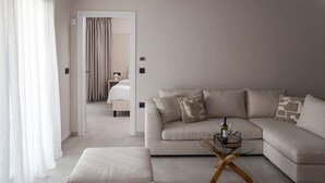 A modern living room with a beige sofa, a glass coffee table, and a view into the bedroom through a doorway.