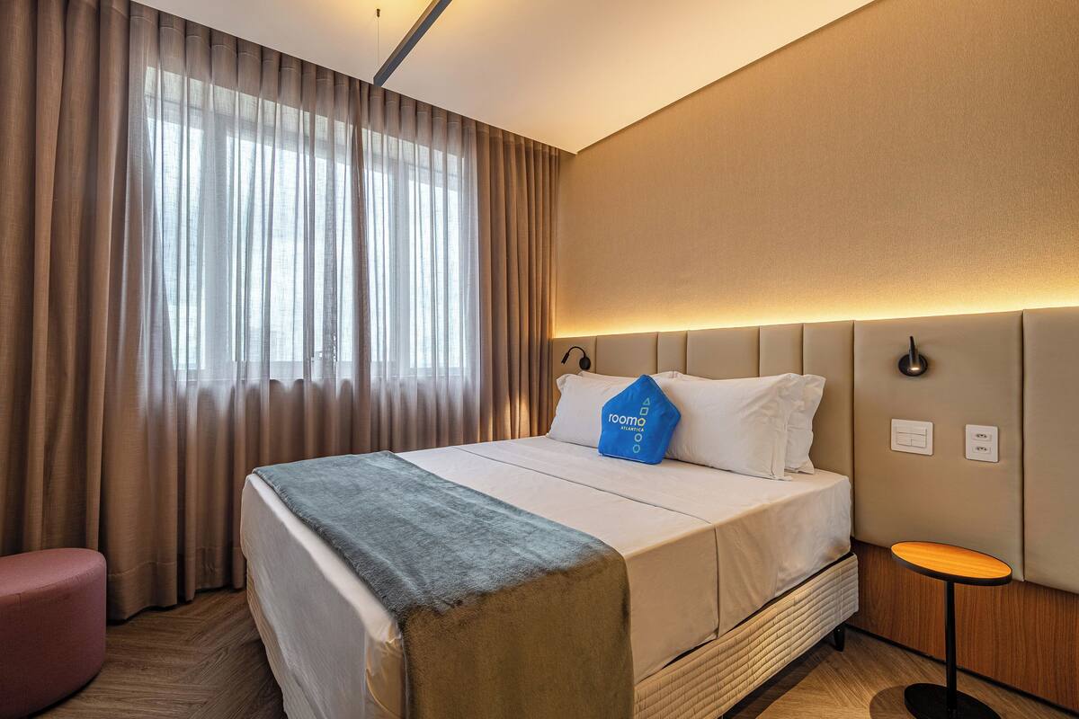 A hotel room with a bed, a bedside table, a chair, and a window with curtains.