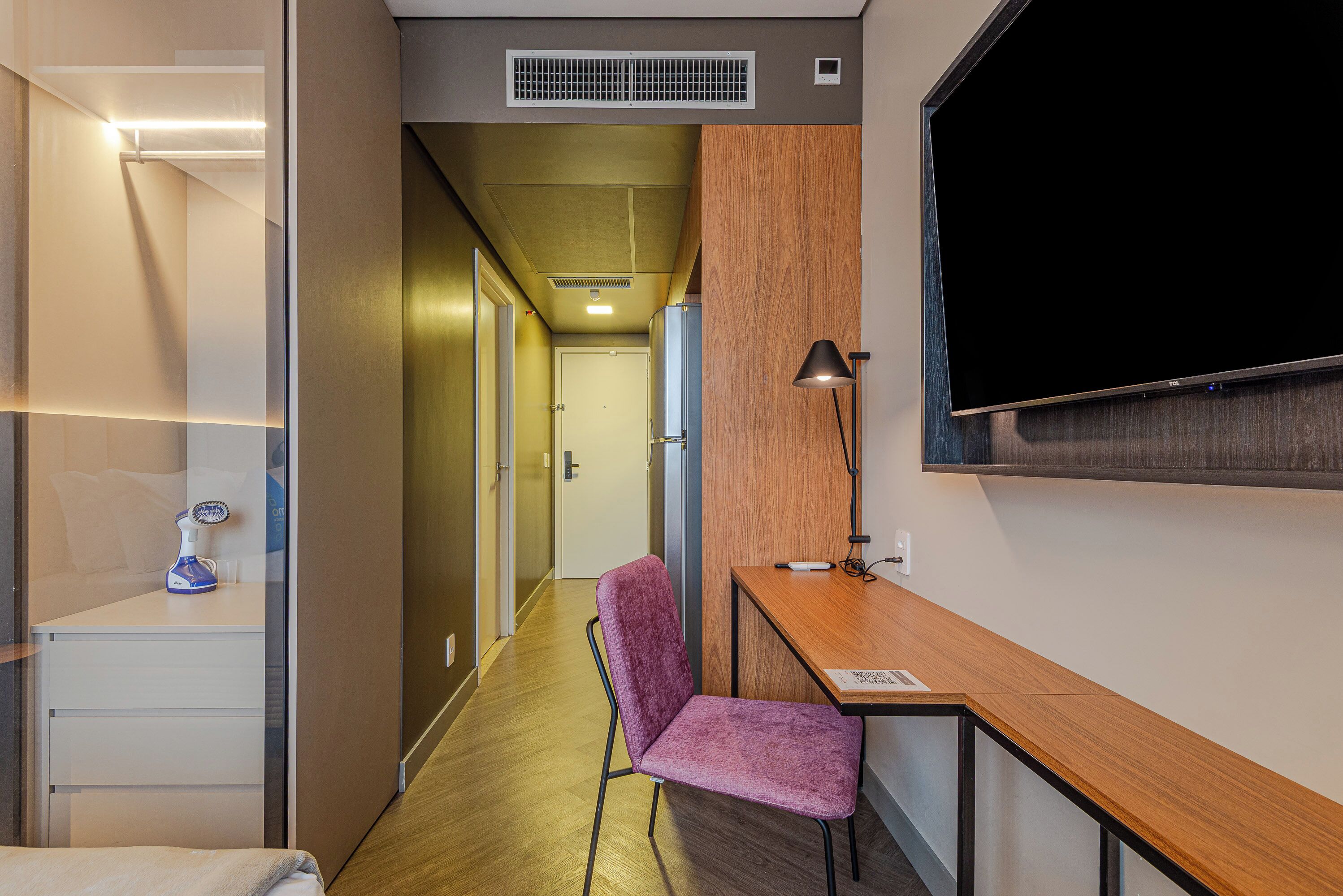 A hotel room with a wooden desk, a chair, a TV, and a lamp.