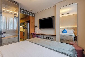 Habitación de hotel con cama, escritorio, silla, televisor y baño con ducha.
