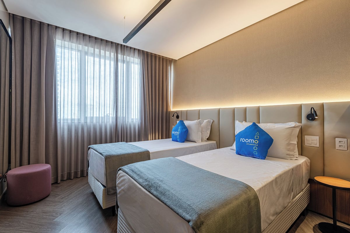 A hotel room with two beds, a small pink ottoman, a bedside table, and a large window with curtains.