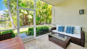 A sunlit room with a glass door leading to a garden, a wicker sofa with cushions, and a wooden table.