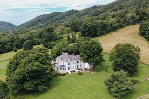A large white house with a symmetrical facade, surrounded by lush green fields and trees, set on a hillside. - Historic Estate w/ 7 KING Suites, Hot Tub, Mountain Views (Hot Springs)
