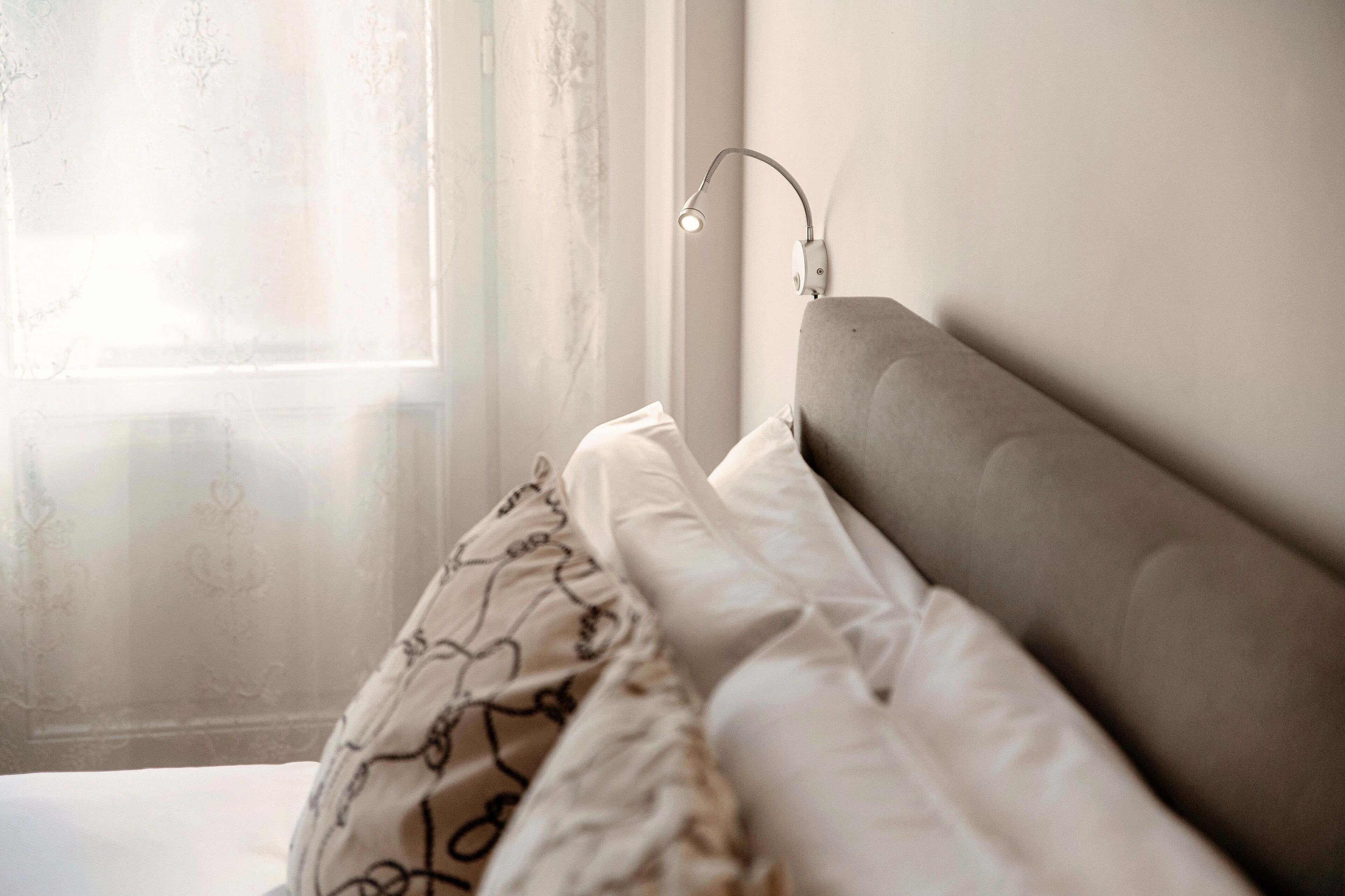 A neatly made bed with a headboard, a bedside lamp, and a window with sheer curtains.