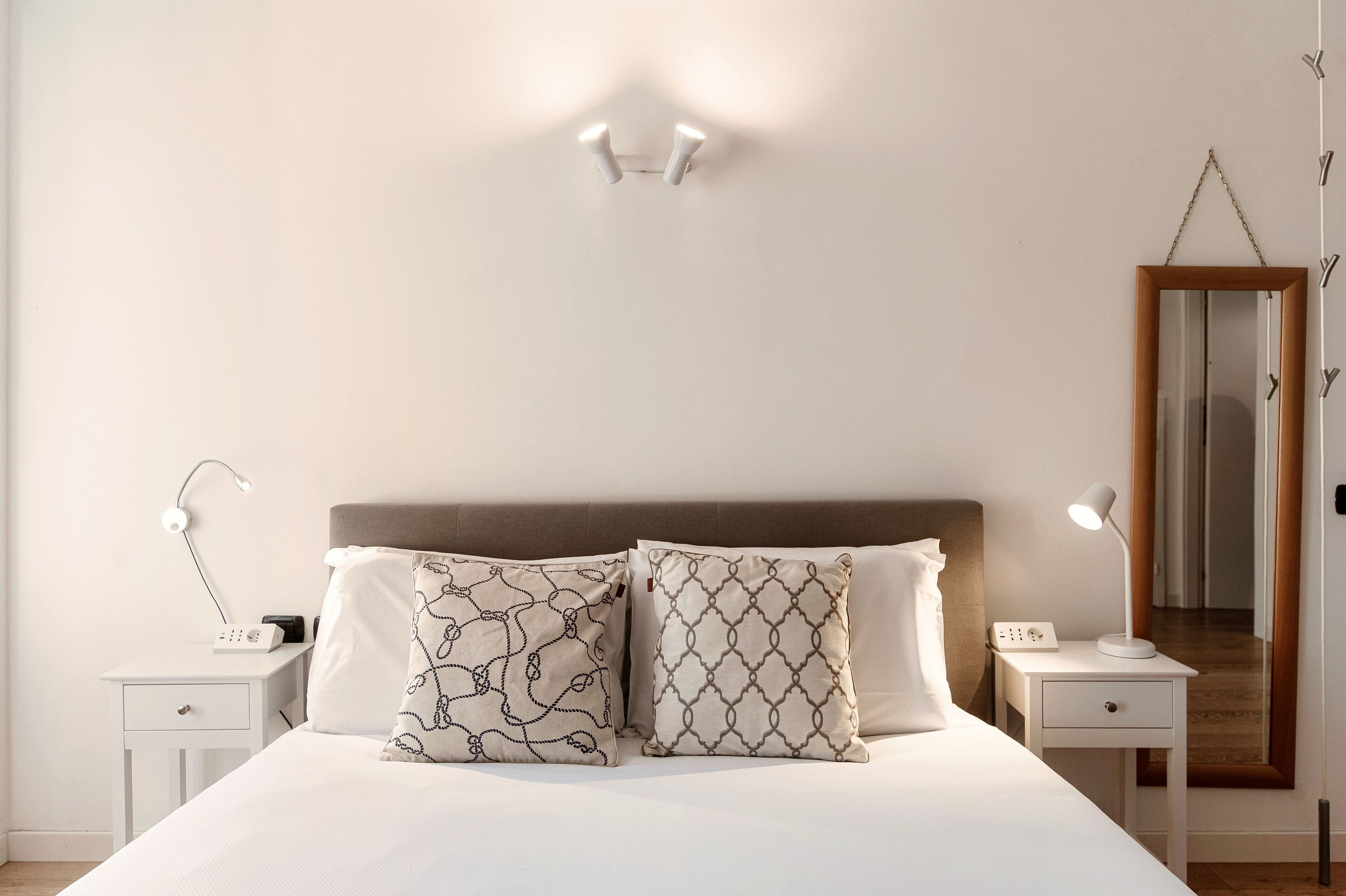 Une chambre à coucher bien rangée, avec un grand lit, deux tables de chevet avec des lampes, une horloge sur la table de chevet et un miroir au mur.